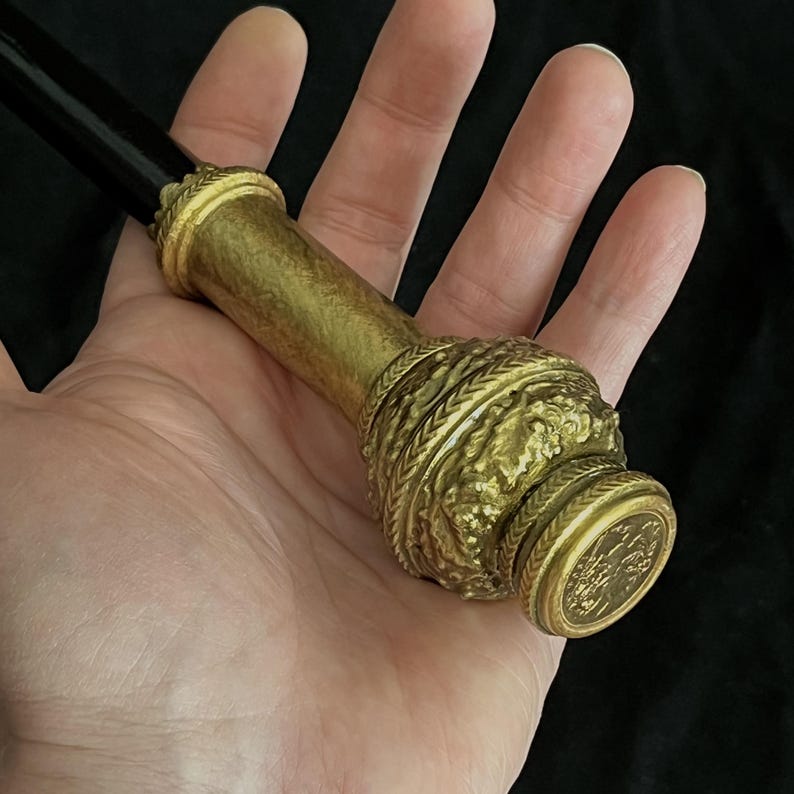 May include: A decorative bottle stopper with a gold-coloured metal finish. The stopper features intricate embossed designs and a black handle. The design includes a detailed pattern around the body and a circular emblem on the end.