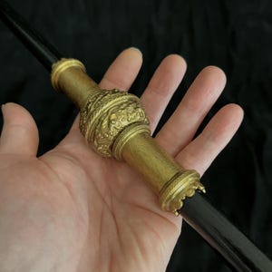 May include: A black wand with elaborate gold-coloured detailing. The wand has a central bulbous section with intricate carvings and decorative bands at each end. The wand is held in a hand against a dark background.