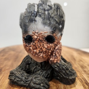 May include: A handcrafted figurine of a character with a tree-like appearance. The figure has black eyes, a copper-coloured face, and a grey and black textured head. The figurine is resting on a wooden surface.
