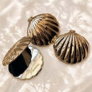 May include: Three gold-colored shell-shaped compact mirrors with a metallic finish. One mirror is open, revealing a reflective surface with the word Mamina engraved in cursive script. The other two mirrors are closed, showing the ribbed shell exterior.