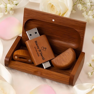May include: A wooden USB flash drive with the names "Stacey & David" and the date "07-09-2025" engraved on it, along with a wedding ring graphic. The USB drive is inside a wooden box with a matching wooden lid and two wooden pieces. The background includes white flowers and pink petals.