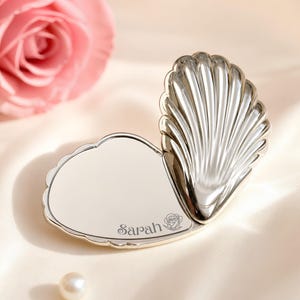 May include: A silver-toned metal compact mirror shaped like an open seashell. The flat surface features the engraved name Sarah next to a small rose illustration. A single white pearl sits nearby, and a pink rose is visible in the upper corner.
