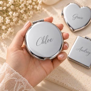 May include: A round silver compact mirror engraved with the name Chloe in a cursive script. Two additional compact mirrors are visible nearby, one heart-shaped with the name Grace and one rectangular with the name Audrey engraved on the surface.