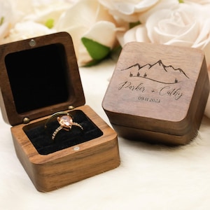 May include: A wooden ring box with a hinged lid, open to reveal a gold ring with a pear-shaped peach-colored stone. The closed box has a mountain design and the names "Parker + Cathy" with the date "09.11.2023" engraved on the lid.