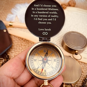 May include: A vintage-style brass compass with a hinged lid, open to reveal a detailed compass face. The lid has an inscription: "And I'd choose you... I'd find you and I'd choose you. Love Sarah." The compass is held in a hand.