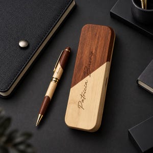May include: A two-toned wooden ballpoint pen and a matching rectangular wooden case. The pen features dark and light wood segments with gold-tone accents and the name Barris engraved. The case is diagonally split with light and dark wood.
