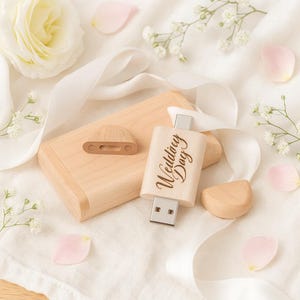 Könnte beinhalten: Ein USB-Stick aus Holz mit der Gravur "Wedding Day", neben einer Holzbox und dekorativen Holzelementen. Das Bild zeigt weißes Band, Blütenblätter und eine weiße Blume, wodurch ein Hochzeitsthema entsteht.