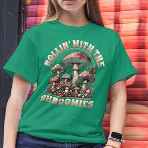 May include: A green t-shirt featuring a mushroom graphic with the text "ROLLIN' WITH THE SHROOMIES." The design includes various mushroom illustrations in shades of red, brown, and blue. The model is wearing blue jeans.