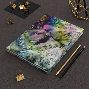 May include: A colorful abstract design notebook with a black spine. The notebook is lying on a gray textured surface with two pencils and a few gold paper clips.