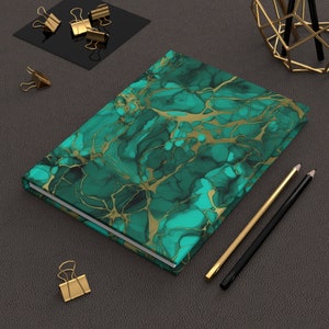 May include: A green and gold marble-patterned notebook with a white border. The notebook is lying on a dark gray surface with gold paper clips and pencils.