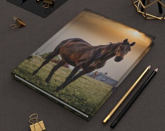 May include: A brown horse stands in a field with a sunset in the background. The horse is looking at the camera. The image is printed on a hardcover journal.