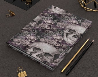 May include: A dark purple journal with a skull and rose pattern. The journal is closed and lying on a gray surface with two pencils and gold paper clips.