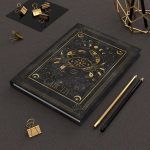 May include: Black journal with gold foil crab zodiac design and the word "CANCER". The cover features celestial elements and a decorative border. Gold pencils and binder clips are on a dark gray surface.