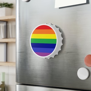 May include: A white bottle opener with a rainbow design. The opener is shaped like a bottle cap and has a magnetic back.