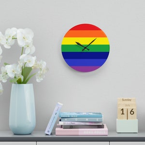 May include: A round clock with a rainbow design. The clock face is white with black hands. The clock is hanging on a white wall. A vase with white flowers is on a shelf below the clock. A wooden calendar block with the date July 16th is on the shelf.