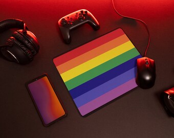 Puede incluir: Un tapete de ratón con colores de arcoíris con un ratón de juego negro, un mando inalámbrico negro, unos auriculares negros y una cámara negra con una lente sobre una superficie oscura.