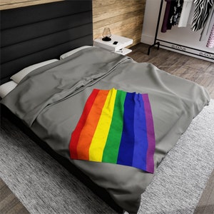 May include: A gray bedspread with a rainbow-colored throw blanket on top. The throw blanket is folded in half and has the colors of the rainbow flag.
