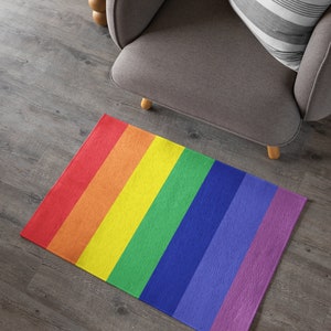 Puede incluir: Una alfombra rectangular con un diseño de arcoíris. La alfombra presenta rayas verticales en los colores del arcoíris: rojo, naranja, amarillo, verde, azul, añil y violeta. La alfombra está colocada sobre un suelo de madera.