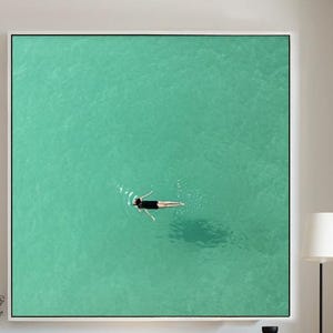 May include: Large square framed art print featuring a swimmer in turquoise water. The artwork hangs above a wooden bench with black metal legs. A black vase with dried plants and other decorative items are on the bench. The room has light wood floors and white walls.