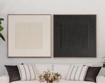 Puede incluir: Dos pinturas abstractas enmarcadas en negro con líneas verticales, una con líneas blancas sobre un fondo beige y la otra con líneas negras sobre un fondo negro, colgadas sobre un sofá blanco con almohadas a rayas blancas y negras, una mesa de centro negra con un jarrón de flores y un puf blanco.