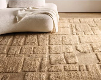 May include: A beige rug with a geometric pattern of raised rectangular shapes. The rug is placed on a wooden floor, with a white sofa and a cream-colored throw blanket in the background. The rug's texture appears soft and plush.