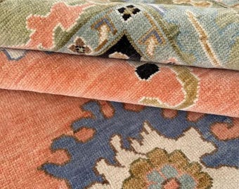 May include: Close-up of a folded rug with a detailed floral pattern. The rug features a color palette of coral, blue, green, and beige. The design includes stylized floral and geometric elements, creating a textured, layered appearance.
