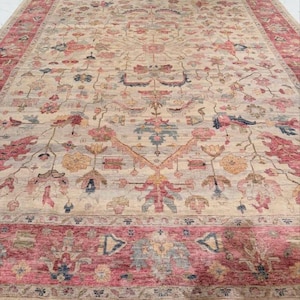 Op de afbeelding: Een groot, rechthoekig vloerkleed met een ingewikkeld bloemmotief. Het vloerkleed heeft een crèmekleurig middenveld met een herhalend ontwerp in roze, blauw en goud. Een brede rand in een dieproze kleur omlijst het vloerkleed.