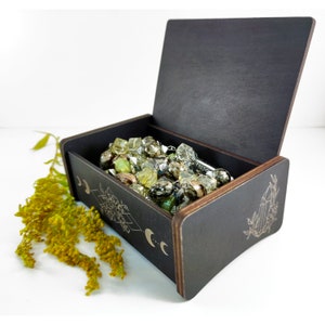 May include: A black wooden box with an engraved design of moons and geometric shapes. The box is open and filled with a variety of green, brown, and clear beads.