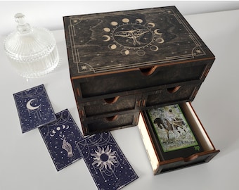 May include: A wooden box with a black stained finish and a decorative design of a moth and moon phases on the lid. The box has five drawers, one of which is open, revealing a tarot card with a depiction of a person riding a horse.