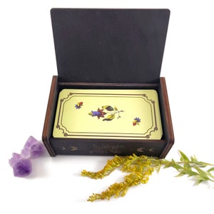 May include: A wooden box with a hinged lid, open to reveal a deck of tarot cards. The box has a dark brown finish and a decorative design on the front. The cards have a yellow background with a floral design in shades of blue, red, and green.