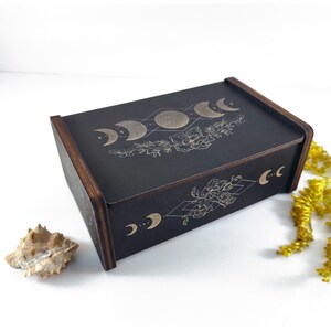 May include: A black wooden box with a gold moon phases design and floral accents. The box has a hinged lid and is perfect for storing small items.