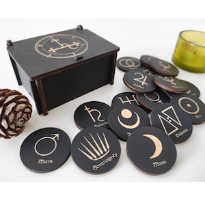 May include: A black wooden box with a lid containing a set of 14 round black wooden tokens with gold symbols and text. The tokens are arranged in a circle around the box. The box has a gold symbol on the lid.