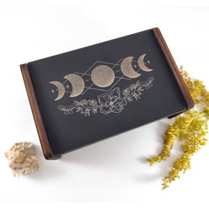 Peut inclure: Une boîte en bois noire avec un motif de phases lunaires dorées et des accents floraux. La boîte a un cadre en bois foncé et une surface peinte en noir.