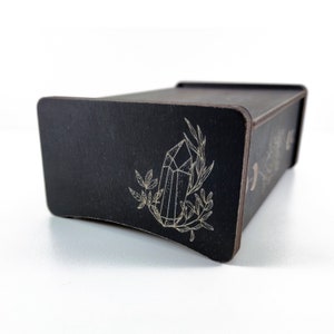 May include: Black wooden box with a drawer, featuring a laser-etched design of a crystal and foliage. The box has a curved front edge.