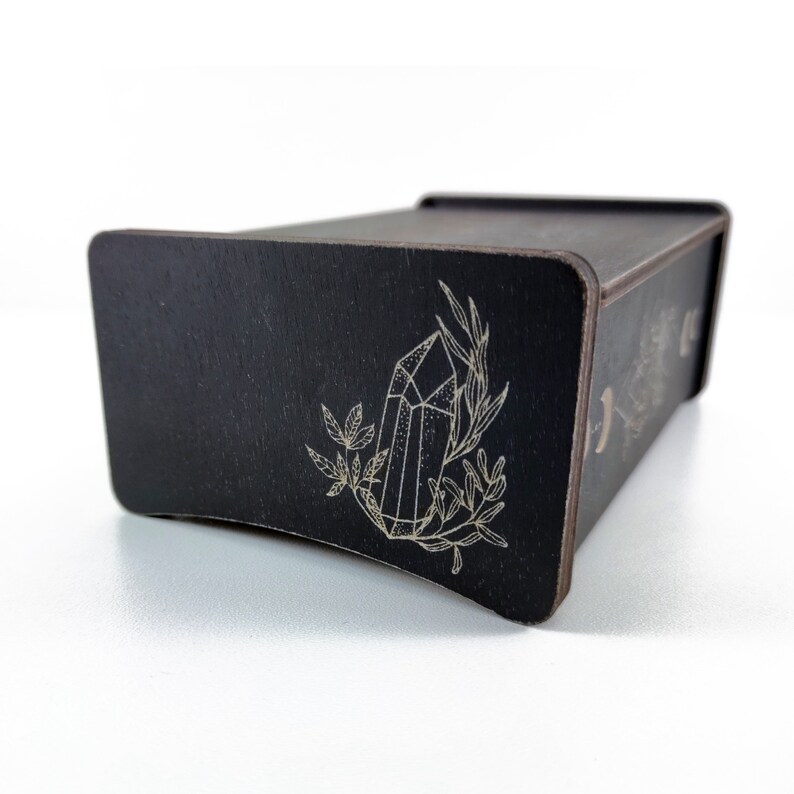 May include: Black wooden box with a drawer, featuring a laser-etched design of a crystal and foliage. The box has a curved front edge.