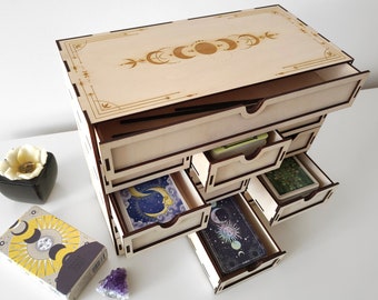 May include: Wooden box with six drawers, each drawer is open and contains a deck of tarot cards. The box has a lid with a moon and star design.