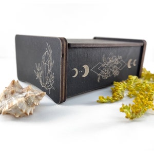 May include: A black wooden box with a geometric design featuring moons, diamonds, and floral elements. The box has a crystal design on the side.