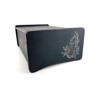 May include: A black wooden box with a crystal and leaves design engraved on the lid. The box is closed and sitting on a white surface.