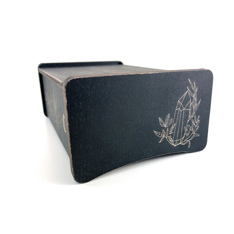 May include: A black wooden box with a crystal and leaves design engraved on the lid. The box is closed and sitting on a white surface.