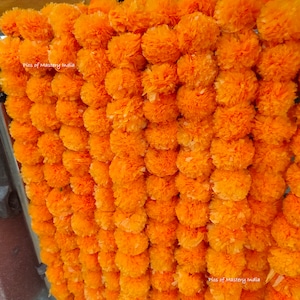 May include: A dense arrangement of bright orange marigold garlands, hanging vertically. The fully bloomed flowers create a textured, layered appearance. The image is well-lit, emphasizing the vivid colour and intricate details of the petals.