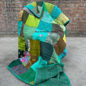 May include: A patchwork quilt in shades of green, teal, and gold. The quilt is made of various fabric pieces stitched together. The text "Pics of Mastery India" is visible on the quilt. The quilt is displayed on a concrete surface against a brick wall.