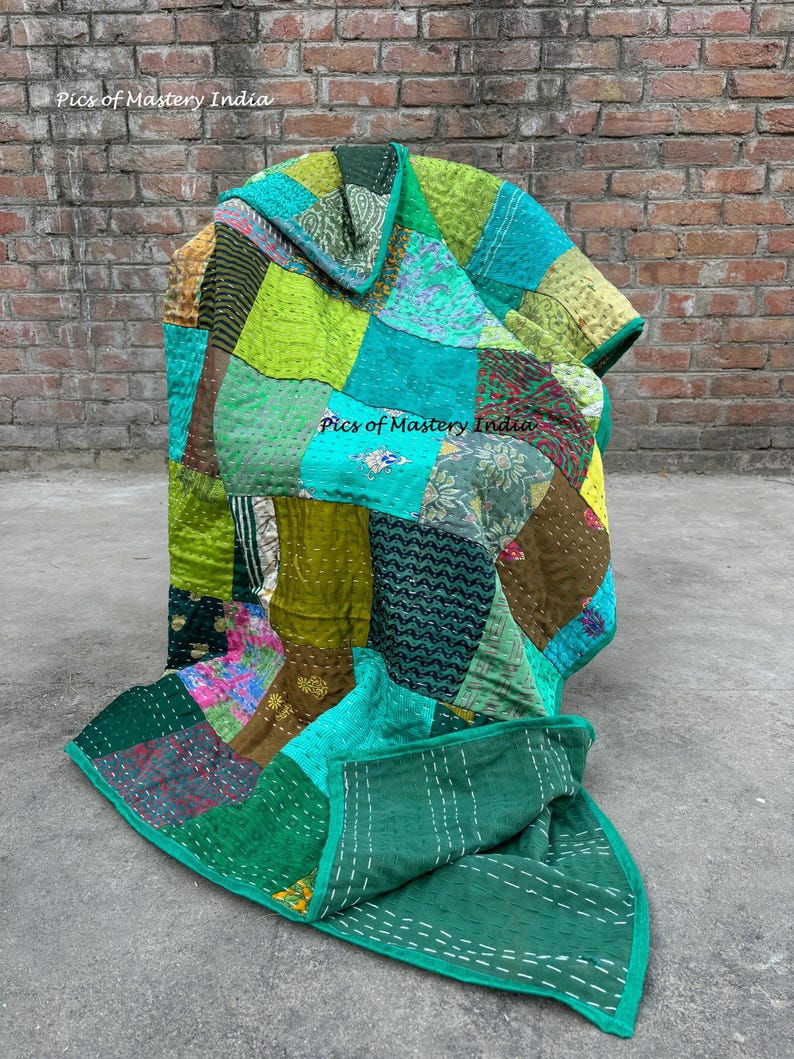 May include: A patchwork quilt in shades of green, teal, and gold. The quilt is made of various fabric pieces stitched together. The text "Pics of Mastery India" is visible on the quilt. The quilt is displayed on a concrete surface against a brick wall.