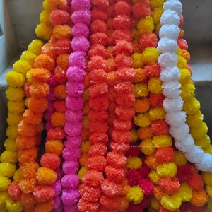 Op de afbeelding: Een verzameling kleurrijke goudsbloemkransen, met tinten oranje, geel, roze en wit. De kransen zijn gemaakt van papier en zijn verticaal gestapeld. De tekst "Pics of Mastery India" is zichtbaar op twee van de kransen.