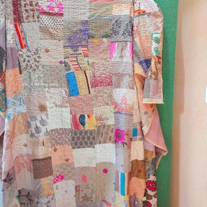 Può includere: Una trapunta patchwork realizzata con una varietà di tessuti in tonalità di marrone, beige, rosa e blu. La trapunta è composta da quadrati di diverse dimensioni e motivi, ed è cucita a mano con un punto a corsa. La trapunta è piegata e appesa a un muro.