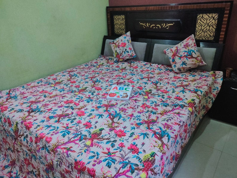 May include: A white bed with a floral patterned duvet cover. The duvet cover features a variety of colorful flowers and birds. The bed has a black headboard with a decorative design. A book with the letters "ABC" is on the bed.