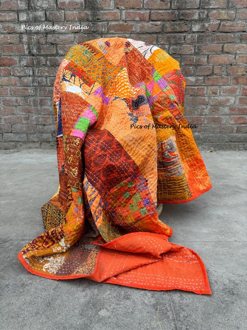 May include: A patchwork quilt in shades of orange, red, and brown, draped over a chair. The quilt features a variety of patterned fabrics and visible stitching. The text "Pics of Mastery India" is visible on the quilt.