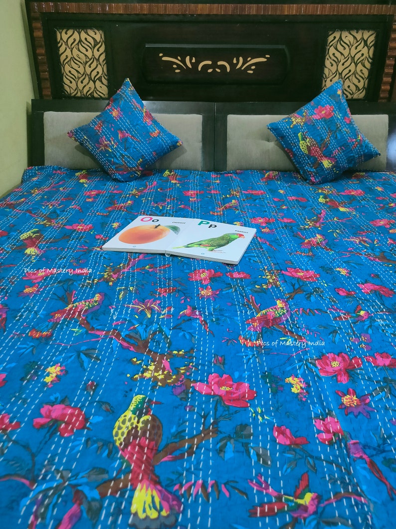May include: A blue kantha quilt with a floral and bird pattern. The quilt is on a bed with two pillows. A children's book with the letters "Oo" and "Pp" is on the quilt.