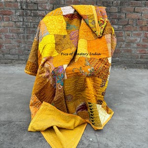 May include: A patchwork quilt in shades of yellow, orange, and brown, draped over a chair. The quilt features a variety of patterns and textures, with visible stitching. The words "Pics of Mastery India" are visible on the quilt.