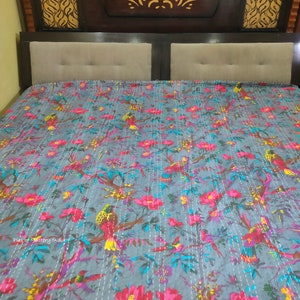 May include: A gray kantha quilt with a floral and bird print. The quilt is made of cotton and has a hand-stitched design. The quilt is shown on a bed with a wooden headboard.