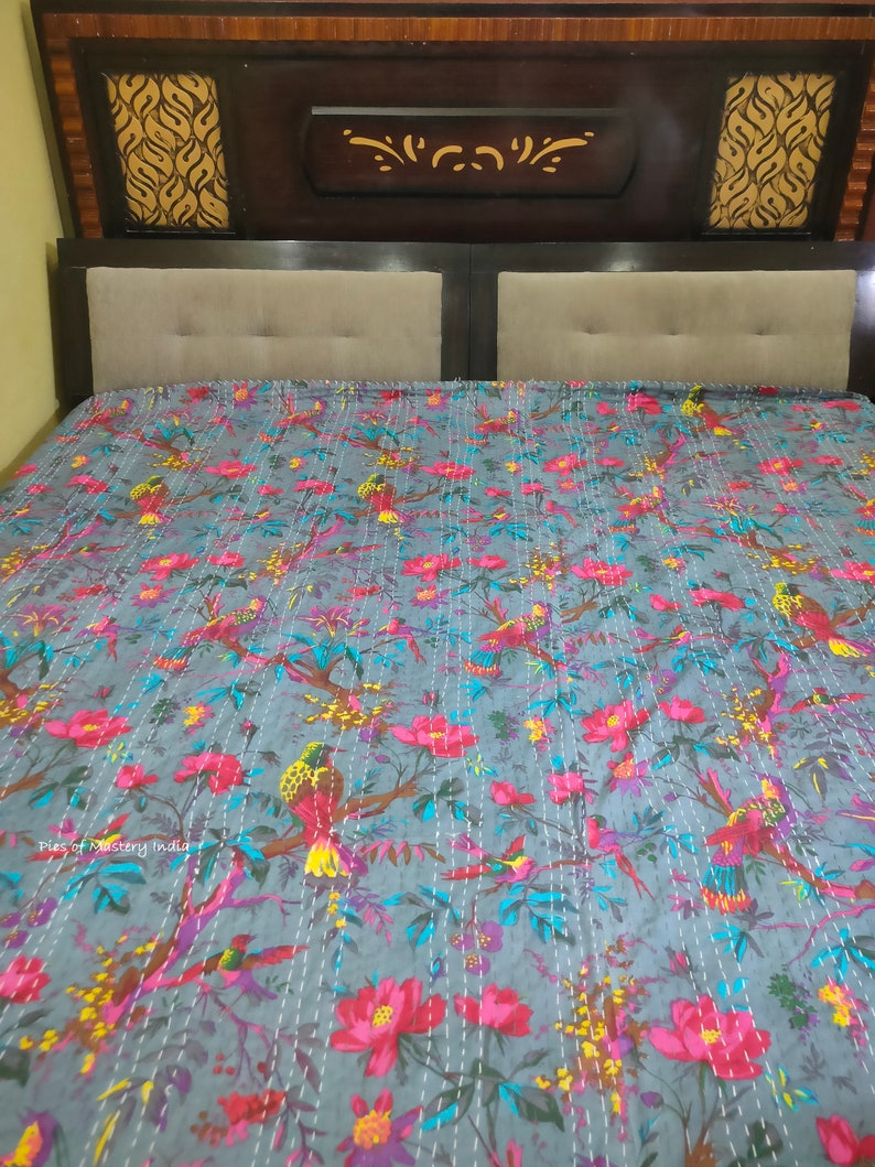 May include: A gray kantha quilt with a floral and bird print. The quilt is made of cotton and has a hand-stitched design. The quilt is shown on a bed with a wooden headboard.